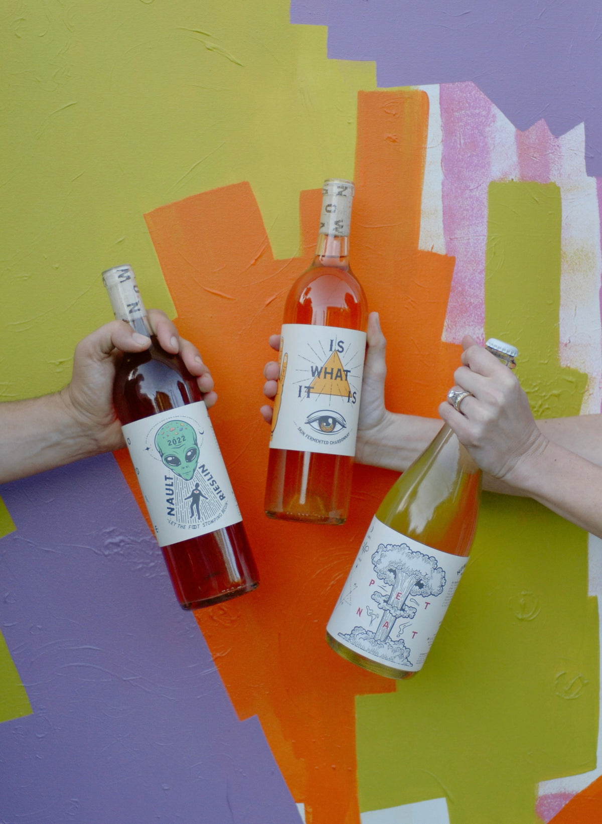 Hands holding three Nowadays wine bottles against a colorful mural