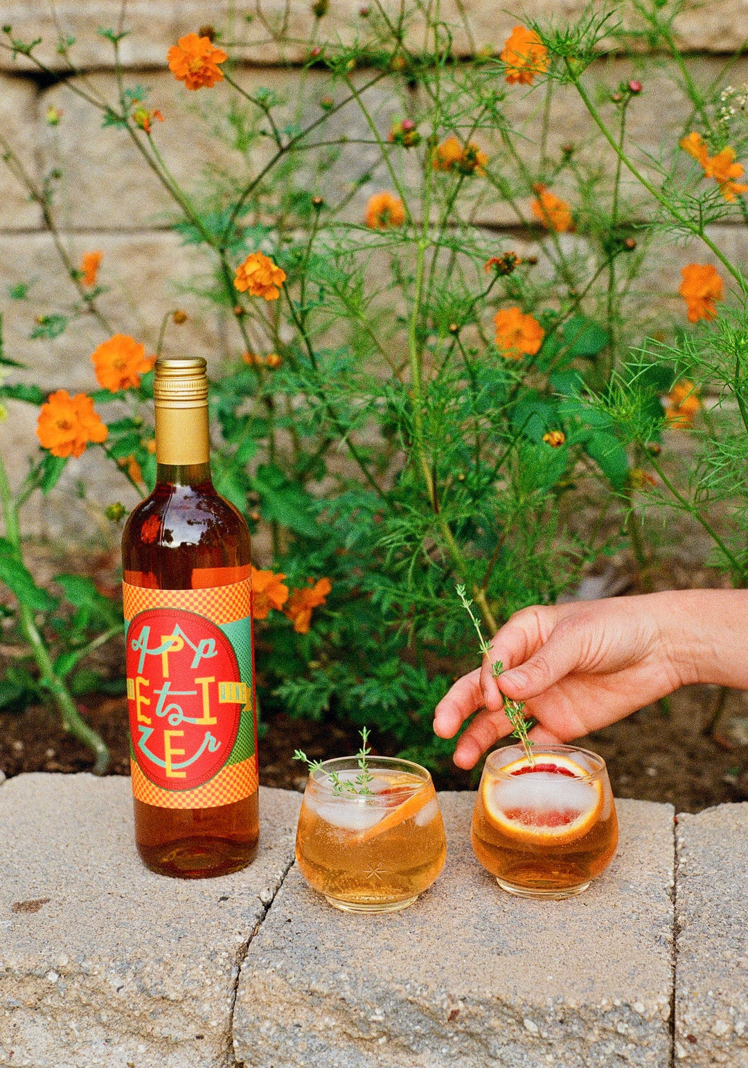 Muni Appetizer aperitif beside two iced citrus drinks, with orange flowers and a hand adding a garnish.