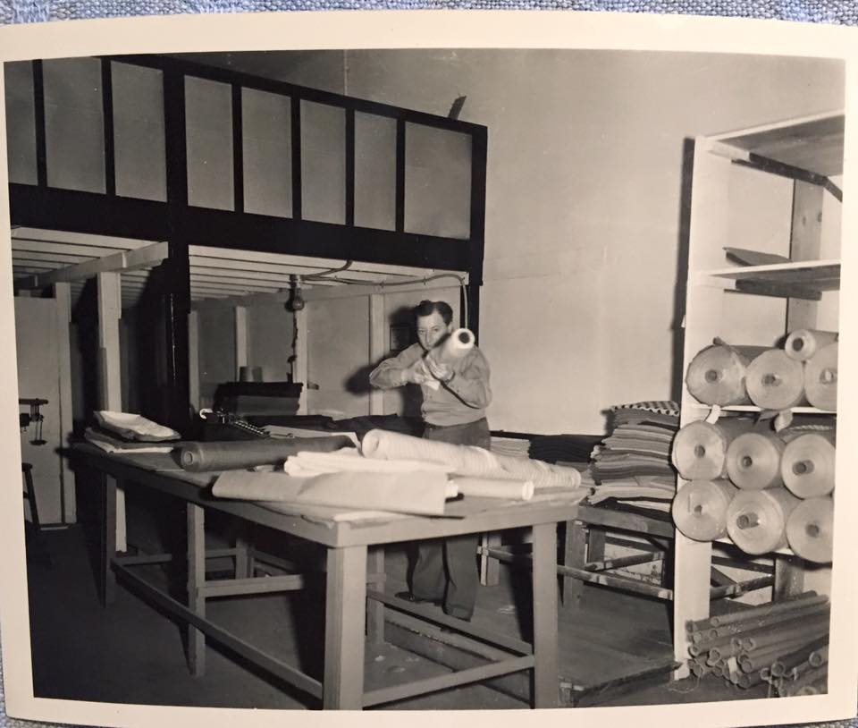 A person holding a bolt of fabric beside a cutting table and shelves of cloth.